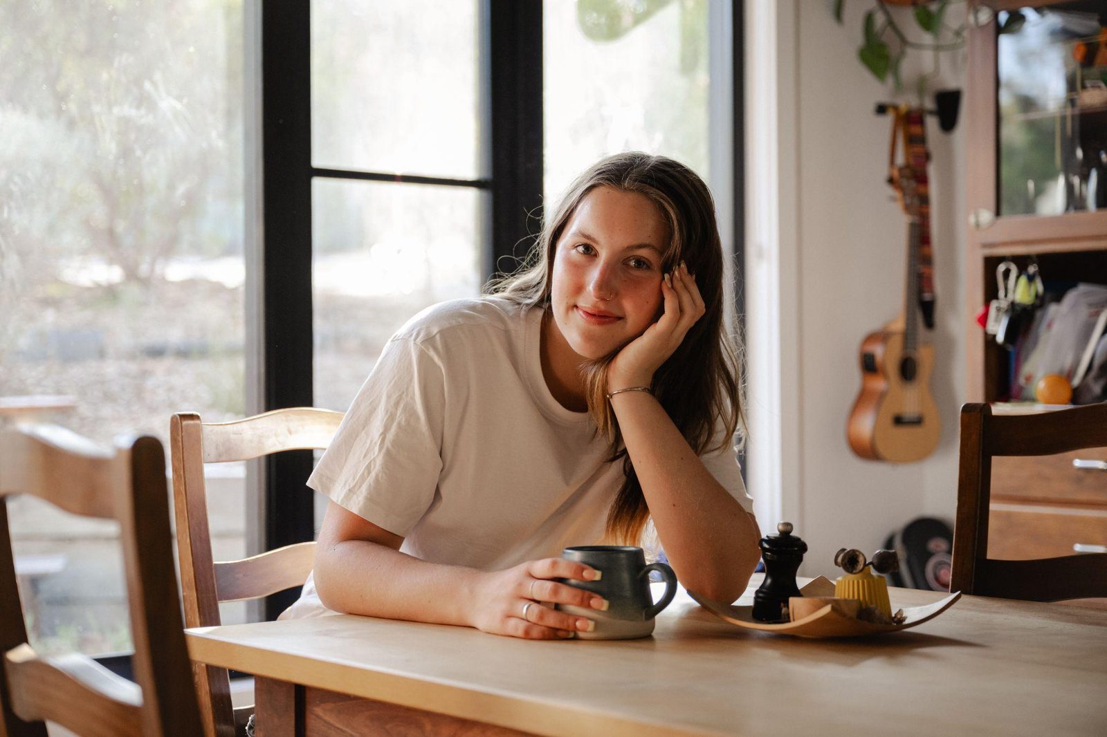 A young woman sitting at a kitchen table by a window, holding a mug